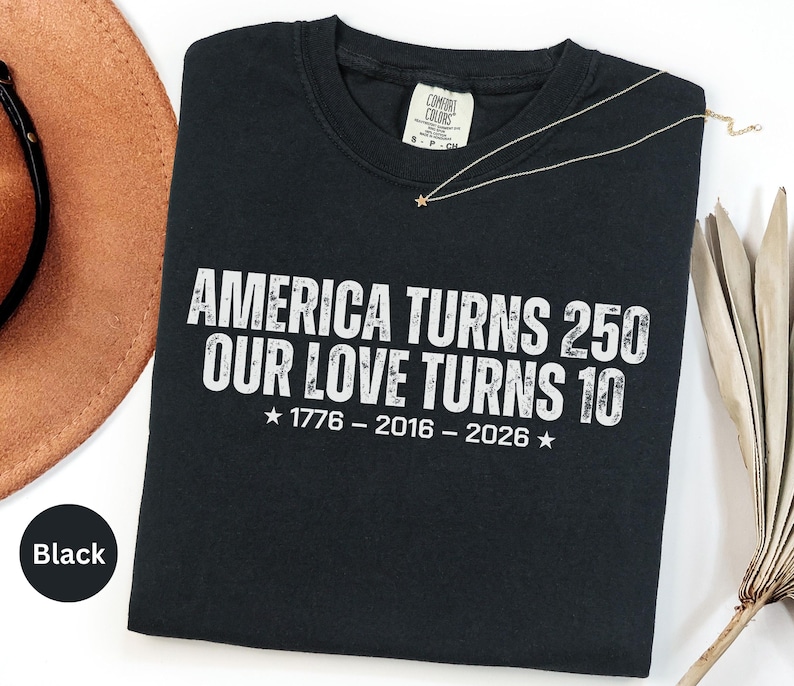 May include: A black short-sleeved t-shirt featuring white distressed text that reads AMERICA TURNS 250 OUR LOVE TURNS 10, with the years 1776 - 2016 - 2026 and two stars printed below. The shirt has a Comfort Colors brand label at the neck.