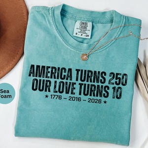 May include: A sea foam green short-sleeve t-shirt with black text that reads: AMERICA TURNS 250 OUR LOVE TURNS 10 1776 - 2016 - 2026. The shirt features a crew neckline and a Comfort Colors brand label on the inside collar, with a gold-toned necklace.