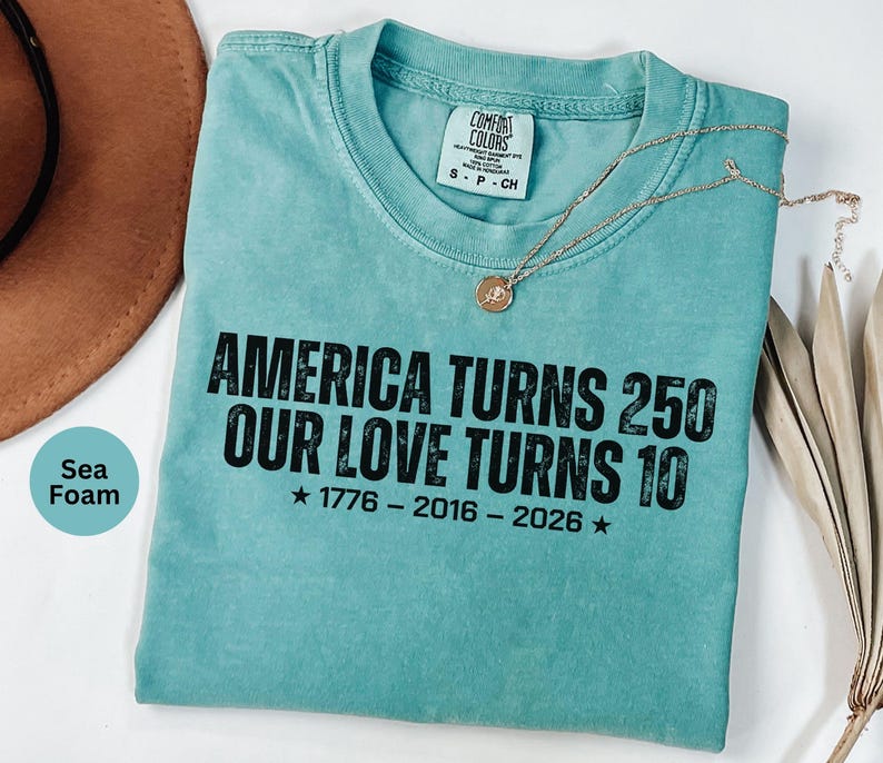 May include: A sea foam green short-sleeve t-shirt with black text that reads: AMERICA TURNS 250 OUR LOVE TURNS 10 1776 - 2016 - 2026. The shirt features a crew neckline and a Comfort Colors brand label on the inside collar, with a gold-toned necklace.