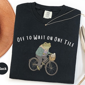 Op de afbeelding: Een zwart t-shirt met een afbeelding van een kikker in een lichtgroen vest en een sjaaltje met patroon, rijdend op een fiets met een mandje met spullen. De tekst "OFF TO WAIT ON ONE TILE" staat in het wit boven de illustratie van de kikker.