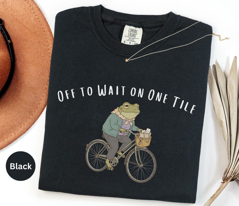 Op de afbeelding: Een zwart t-shirt met een afbeelding van een kikker in een lichtgroen vest en een sjaaltje met patroon, rijdend op een fiets met een mandje met spullen. De tekst "OFF TO WAIT ON ONE TILE" staat in het wit boven de illustratie van de kikker.