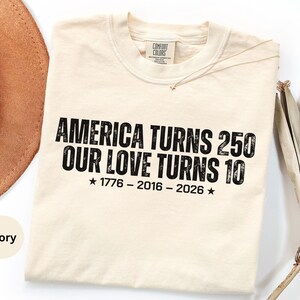 May include: An ivory-colored crew neck t-shirt featuring black distressed-style text that reads: AMERICA TURNS 250 OUR LOVE TURNS 10, followed by the years 1776 - 2016 - 2026 with a star on each side. The shirt has a Comfort Colors brand neck label.