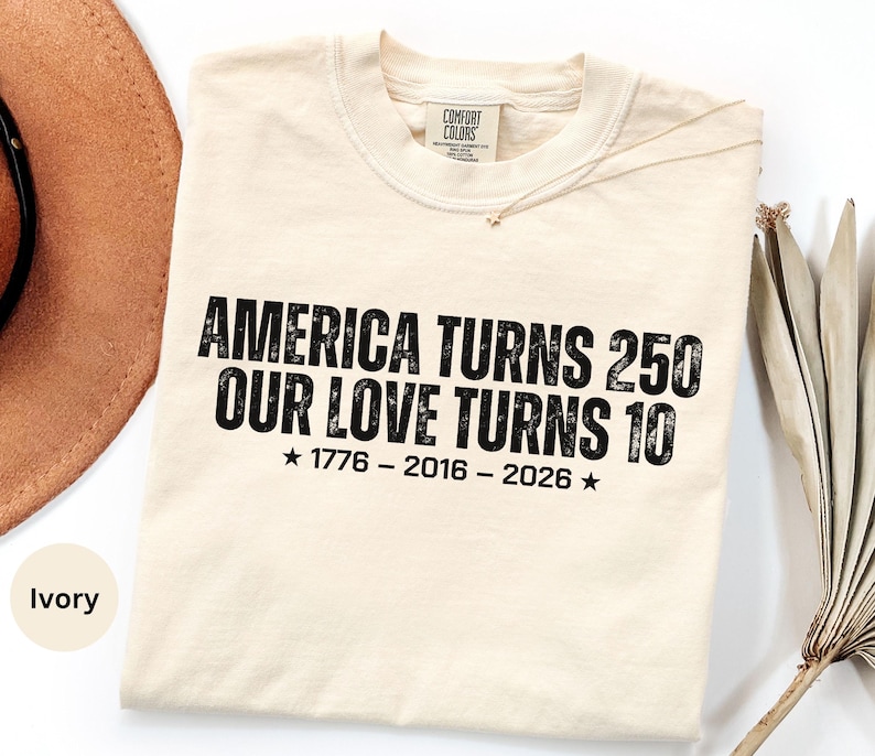 May include: An ivory-colored crew neck t-shirt featuring black distressed-style text that reads: AMERICA TURNS 250 OUR LOVE TURNS 10, followed by the years 1776 - 2016 - 2026 with a star on each side. The shirt has a Comfort Colors brand neck label.