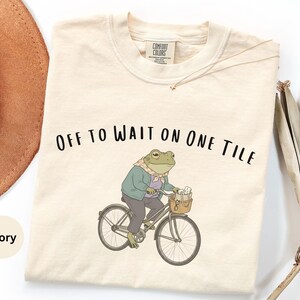 Op de afbeelding: Een ivoorkleurig t-shirt met een afbeelding van een kikker in een jasje en sjaal op een fiets. De kikker heeft een mandje aan het stuur met voorwerpen erin. De tekst "OFF TO WAIT ON ONE TILE" staat in het zwart boven de illustratie gedrukt.