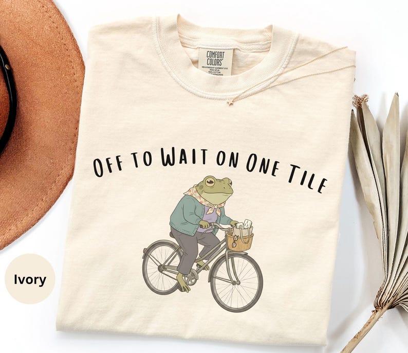 Op de afbeelding: Een ivoorkleurig t-shirt met een afbeelding van een kikker in een jasje en sjaal op een fiets. De kikker heeft een mandje aan het stuur met voorwerpen erin. De tekst "OFF TO WAIT ON ONE TILE" staat in het zwart boven de illustratie gedrukt.
