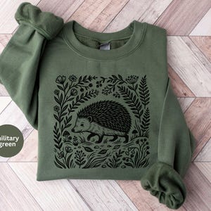 Peut inclure: Un sweat-shirt à col rond vert militaire avec une impression graphique noire représentant un hérisson entouré de diverses plantes, fleurs et petits champignons. Le motif est centré sur le devant du vêtement, doté de manches longues et de poignets côtelés.
