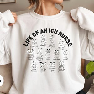 May include: White sweatshirt with the text "LIFE OF AN ICU NURSE" in black. The design includes various cartoon faces with expressions and phrases like "code already?" and "night shift things."