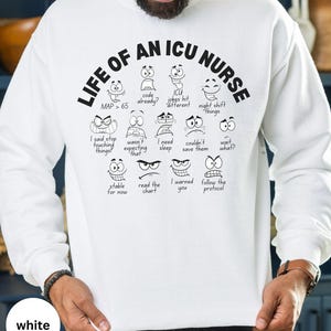 May include: White sweatshirt with the text "LIFE OF AN ICU NURSE" in black, featuring various cartoon faces expressing different emotions. The sweatshirt also includes phrases like "MAP > 65" and "code already?"