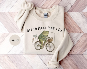 May include: A sand-colored crewneck sweatshirt featuring a graphic of a green frog wearing a stethoscope and riding a bicycle. The frog has a basket with a clipboard and a hospital sign in the background. The text reads OFF TO MAKE MAP > 65.