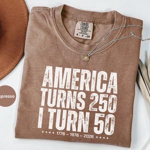 May include: Espresso-colored t-shirt with the text "AMERICA TURNS 250 I TURN 50" in white block letters. The shirt also features the years 1776-1976-2026. A silver necklace with a pendant rests on the shirt.
