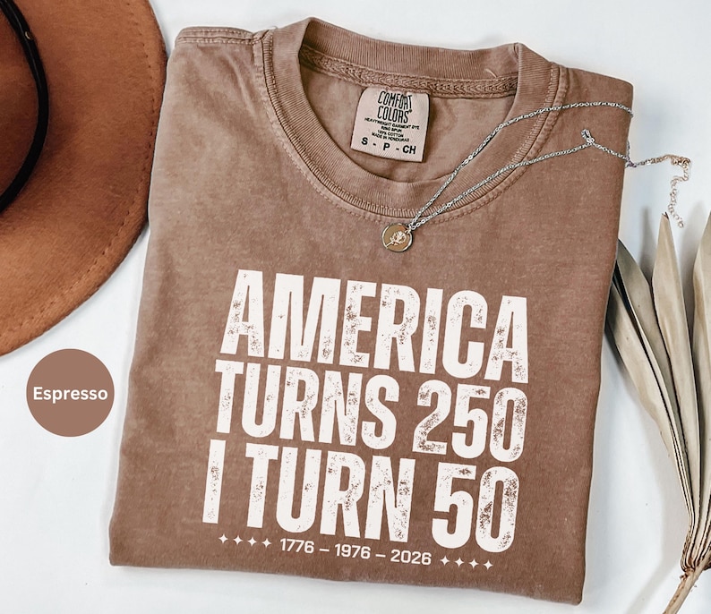 May include: Espresso-colored t-shirt with the text "AMERICA TURNS 250 I TURN 50" in white block letters. The shirt also features the years 1776-1976-2026. A silver necklace with a pendant rests on the shirt.