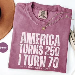 May include: Berry-colored t-shirt with the text "AMERICA TURNS 250 I TURN 70" in white block letters. The shirt includes the years 1776-1956-2026. A gold necklace and a brown hat are visible.