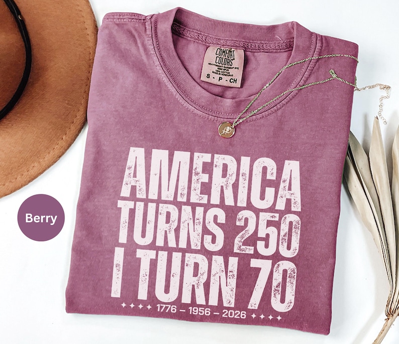 May include: Berry-colored t-shirt with the text "AMERICA TURNS 250 I TURN 70" in white block letters. The shirt includes the years 1776-1956-2026. A gold necklace and a brown hat are visible.