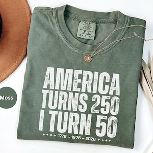 May include: A moss green t-shirt with the text "AMERICA TURNS 250 I TURN 50" in white. The shirt has a crew neck and short sleeves. A gold necklace with a pendant is on the shirt. The shirt is size S-P-CH.