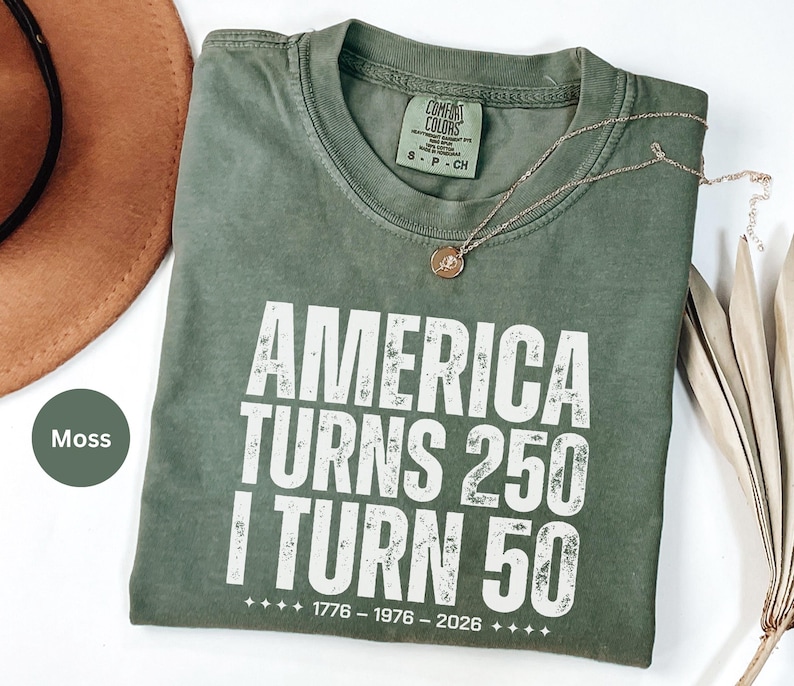 May include: A moss green t-shirt with the text "AMERICA TURNS 250 I TURN 50" in white. The shirt has a crew neck and short sleeves. A gold necklace with a pendant is on the shirt. The shirt is size S-P-CH.