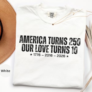 May include: A white short-sleeved t-shirt featuring black distressed text that reads AMERICA TURNS 250 OUR LOVE TURNS 10 with the dates 1776 - 2016 - 2026 and two stars. The shirt has a Comfort Colors brand label at the inner neckline.