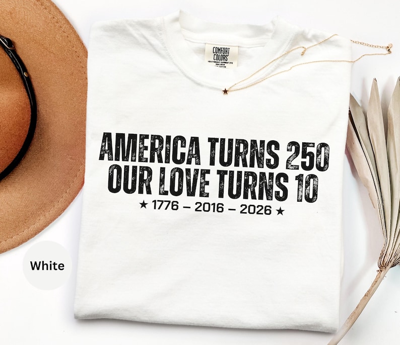 May include: A white short-sleeved t-shirt featuring black distressed text that reads AMERICA TURNS 250 OUR LOVE TURNS 10 with the dates 1776 - 2016 - 2026 and two stars. The shirt has a Comfort Colors brand label at the inner neckline.