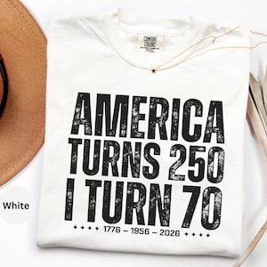 May include: White t-shirt with the text "AMERICA TURNS 250 I TURN 70" in bold black letters. The shirt also includes the dates "1776 - 1956 - 2026" below the text. A gold necklace with a star pendant is visible.