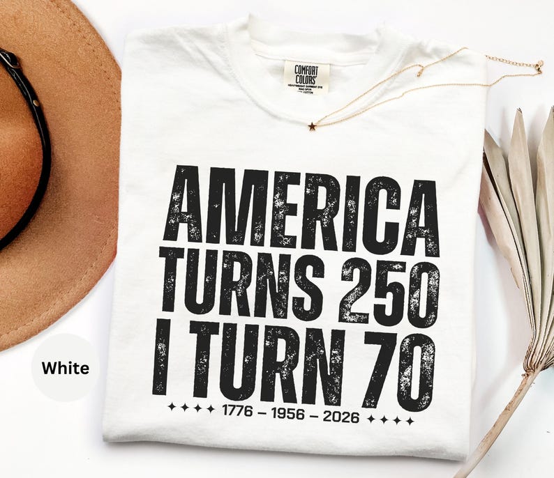 May include: White t-shirt with the text "AMERICA TURNS 250 I TURN 70" in bold black letters. The shirt also includes the dates "1776 - 1956 - 2026" below the text. A gold necklace with a star pendant is visible.