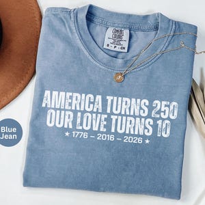 May include: A blue jean colored t-shirt with white text that reads AMERICA TURNS 250 OUR LOVE TURNS 10, with the years 1776 - 2016 - 2026 printed below. The shirt features a crew neckline and a Comfort Colors brand label at the inner collar.