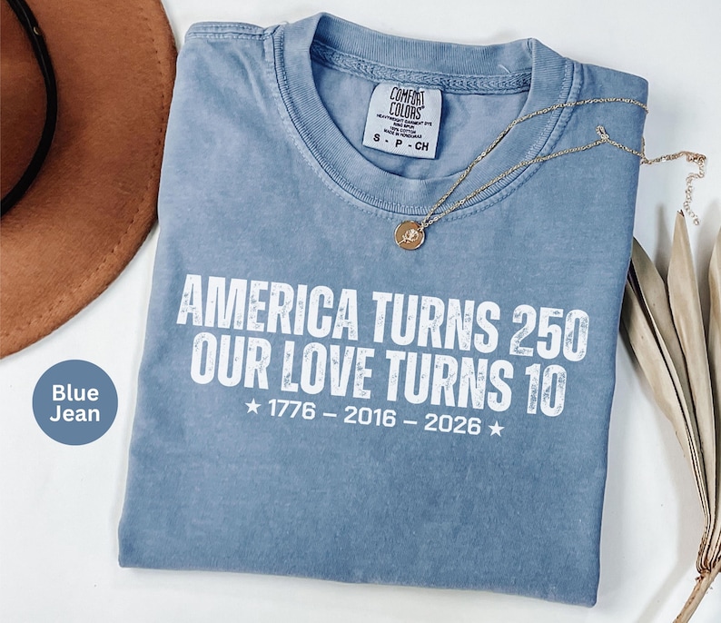 May include: A blue jean colored t-shirt with white text that reads AMERICA TURNS 250 OUR LOVE TURNS 10, with the years 1776 - 2016 - 2026 printed below. The shirt features a crew neckline and a Comfort Colors brand label at the inner collar.