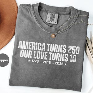 May include: A pepper-colored crew neck t-shirt featuring white distressed text that reads AMERICA TURNS 250 OUR LOVE TURNS 10, with the years 1776 - 2016 - 2026 centered below, accompanied by two stars. The shirt has a Comfort Colors brand label.