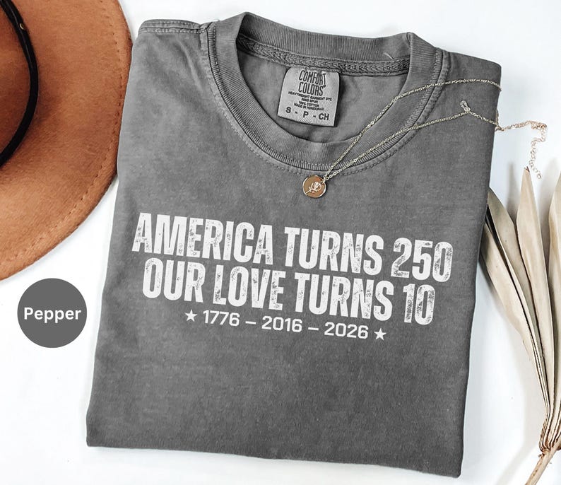 May include: A pepper-colored crew neck t-shirt featuring white distressed text that reads AMERICA TURNS 250 OUR LOVE TURNS 10, with the years 1776 - 2016 - 2026 centered below, accompanied by two stars. The shirt has a Comfort Colors brand label.