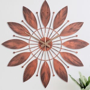 May include: A copper-coloured wall clock with a leaf-shaped design. The clock has a circular face with gold-coloured hands and a matching copper-coloured frame. The clock is mounted on a white wall.