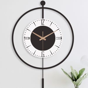 Peut inclure: Une horloge murale moderne avec un cadre circulaire noir et un cadran blanc. Le cadran comporte des chiffres noirs et un centre noir. Les aiguilles de l'horloge sont dorées. Un pendule noir est suspendu en dessous.