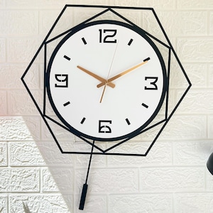 May include: A modern wall clock with a white face, black numerals, and gold-colored hands. The clock is encased in a black geometric frame and has a black pendulum. The clock is hanging on a white brick wall.