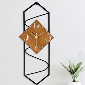 May include: A contemporary wall clock with a geometric design. The clock has a square wooden face with gold hands and a black metal frame. The numbers 12 and 3 are visible. The clock is mounted on a white wall.