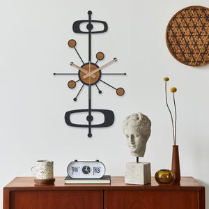 May include: A mid-century modern wall clock with a black frame and wooden accents hangs above a wooden cabinet. The clock features a starburst design with a circular wooden center. Other decor includes a bust, a flip clock, and woven wall hangings.