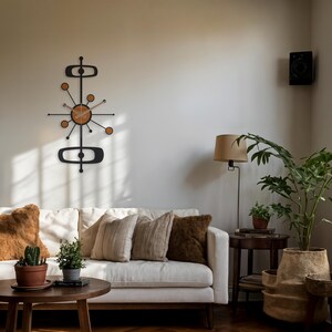 May include: A mid-century modern wall clock with a black metal frame and a light wood-tone clock face. The clock has a unique atomic design with circular and oblong shapes. The clock is mounted on a neutral-colored wall above a white sofa.