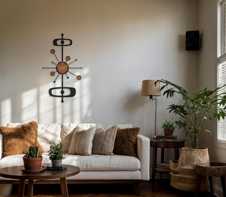 May include: A mid-century modern wall clock with a black metal frame and a light wood-tone clock face. The clock has a unique atomic design with circular and oblong shapes. The clock is mounted on a neutral-colored wall above a white sofa.