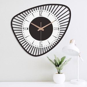 K&ouml;nnte beinhalten: Eine moderne Wanduhr mit schwarzem Rahmen und wei&szlig;em Zifferblatt. Die Uhr hat goldfarbene Zeiger und eine schwarze Mitte. Der Rahmen hat eine unregelm&auml;&szlig;ige Form mit vertikalen Linien, die vom Zifferblatt ausgehen. Die Uhr ist an einer wei&szlig;en Wand befestigt.