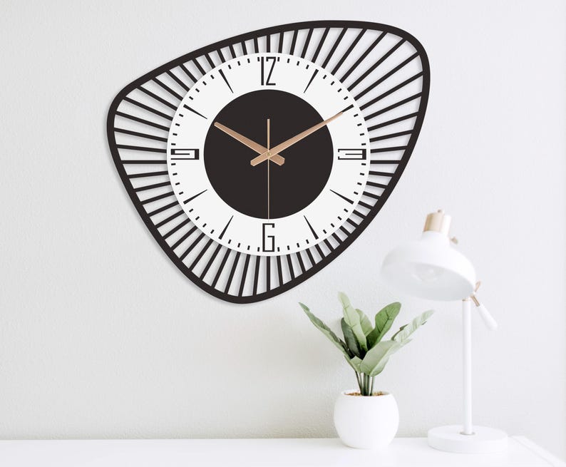 K&ouml;nnte beinhalten: Eine moderne Wanduhr mit schwarzem Rahmen und wei&szlig;em Zifferblatt. Die Uhr hat goldfarbene Zeiger und eine schwarze Mitte. Der Rahmen hat eine unregelm&auml;&szlig;ige Form mit vertikalen Linien, die vom Zifferblatt ausgehen. Die Uhr ist an einer wei&szlig;en Wand befestigt.