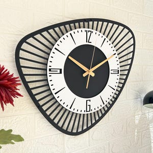 K&ouml;nnte beinhalten: Eine moderne Wanduhr mit einem schwarzen, geometrischen Rahmen. Das Zifferblatt ist wei&szlig; mit schwarzen Ziffern und einem schwarzen Zentrum. Goldfarbene Zeiger zeigen die Uhrzeit an. Die Uhr ist an einer wei&szlig;en Backsteinmauer befestigt.