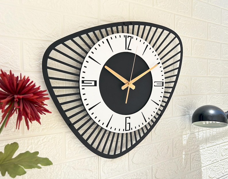 K&ouml;nnte beinhalten: Eine moderne Wanduhr mit einem schwarzen, geometrischen Rahmen. Das Zifferblatt ist wei&szlig; mit schwarzen Ziffern und einem schwarzen Zentrum. Goldfarbene Zeiger zeigen die Uhrzeit an. Die Uhr ist an einer wei&szlig;en Backsteinmauer befestigt.