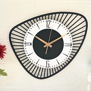 K&ouml;nnte beinhalten: Eine moderne Wanduhr mit einem schwarzen, geometrischen Rahmen und einem wei&szlig;en und schwarzen Zifferblatt. Die Uhr hat goldfarbene Zeiger und ein minimalistisches Design. Die Uhr ist an einer wei&szlig;en Backsteinmauer befestigt.