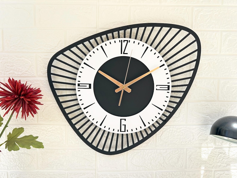 K&ouml;nnte beinhalten: Eine moderne Wanduhr mit einem schwarzen, geometrischen Rahmen und einem wei&szlig;en und schwarzen Zifferblatt. Die Uhr hat goldfarbene Zeiger und ein minimalistisches Design. Die Uhr ist an einer wei&szlig;en Backsteinmauer befestigt.