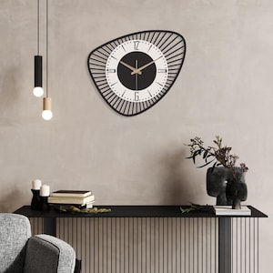 K&ouml;nnte beinhalten: Eine moderne Wanduhr in Schwarz und Wei&szlig; mit einer einzigartigen, asymmetrischen Form. Das Zifferblatt hat eine schwarze Mitte mit wei&szlig;en Ziffern und Zeigern. Die Uhr ist an einer neutralfarbenen Wand montiert, darunter dekorative Elemente.