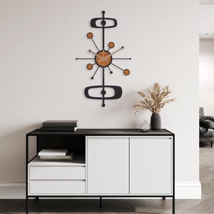 May include: A mid-century modern wall clock with a black frame and wooden accents. The clock features a circular face with radiating arms and circular details. The clock is mounted on a white wall above a black and white cabinet.