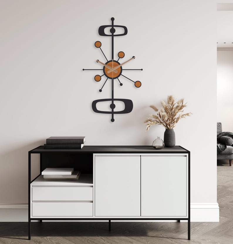 May include: A mid-century modern wall clock with a black frame and wooden accents. The clock features a circular face with radiating arms and circular details. The clock is mounted on a white wall above a black and white cabinet.