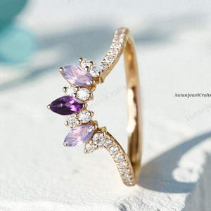 May include: A gold-colored ring with a curved design. The ring features a cluster of marquise-cut purple and lavender gemstones, accented with small white gemstones. The band is encrusted with small white gemstones.