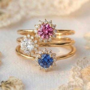 Peut inclure: Trois bagues en or avec des pierres précieuses au centre. Les bagues présentent une pierre rose, blanche et bleue, chacune entourée d'un halo de petites pierres. Les bagues sont empilées les unes sur les autres.