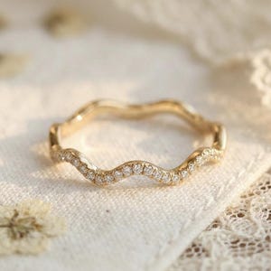 Könnte beinhalten: Ein goldfarbener Ring mit wellenförmigem Design, besetzt mit kleinen, funkelnden Edelsteinen. Der Ring wird auf einem weißen Tuch mit Trockenblumen und Spitze im Hintergrund präsentiert. Das Design des Rings erinnert an eine Welle.