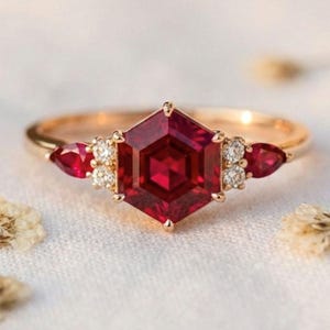 May include: A gold ring with a large, hexagonal, deep red gemstone at its centre. Two smaller, pear-shaped red gems flank the central stone, with small white accent stones. The ring is set against a soft, white background.