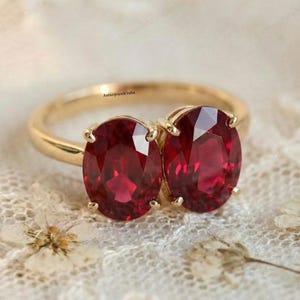 May include: A gold ring featuring two oval-cut, deep red gemstones. The ring's band is a polished gold, and the gemstones are set with four prongs. The ring is displayed on a delicate, off-white lace surface.