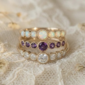Puede incluir: Tres anillos de oro apilados. El anillo superior tiene piedras de ópalo blanco, el anillo central tiene piedras de amatista púrpura y el anillo inferior tiene piedras de diamante transparente. Están sobre encaje.