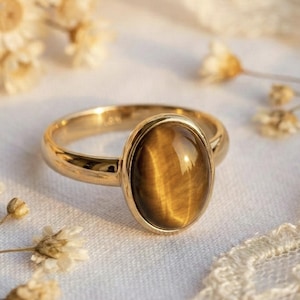 May include: A gold ring featuring an oval tiger's eye gemstone. The ring has a polished gold band and a bezel setting. The tiger's eye stone is a warm brown colour with golden chatoyancy. The ring is displayed on a white surface with dried flowers.